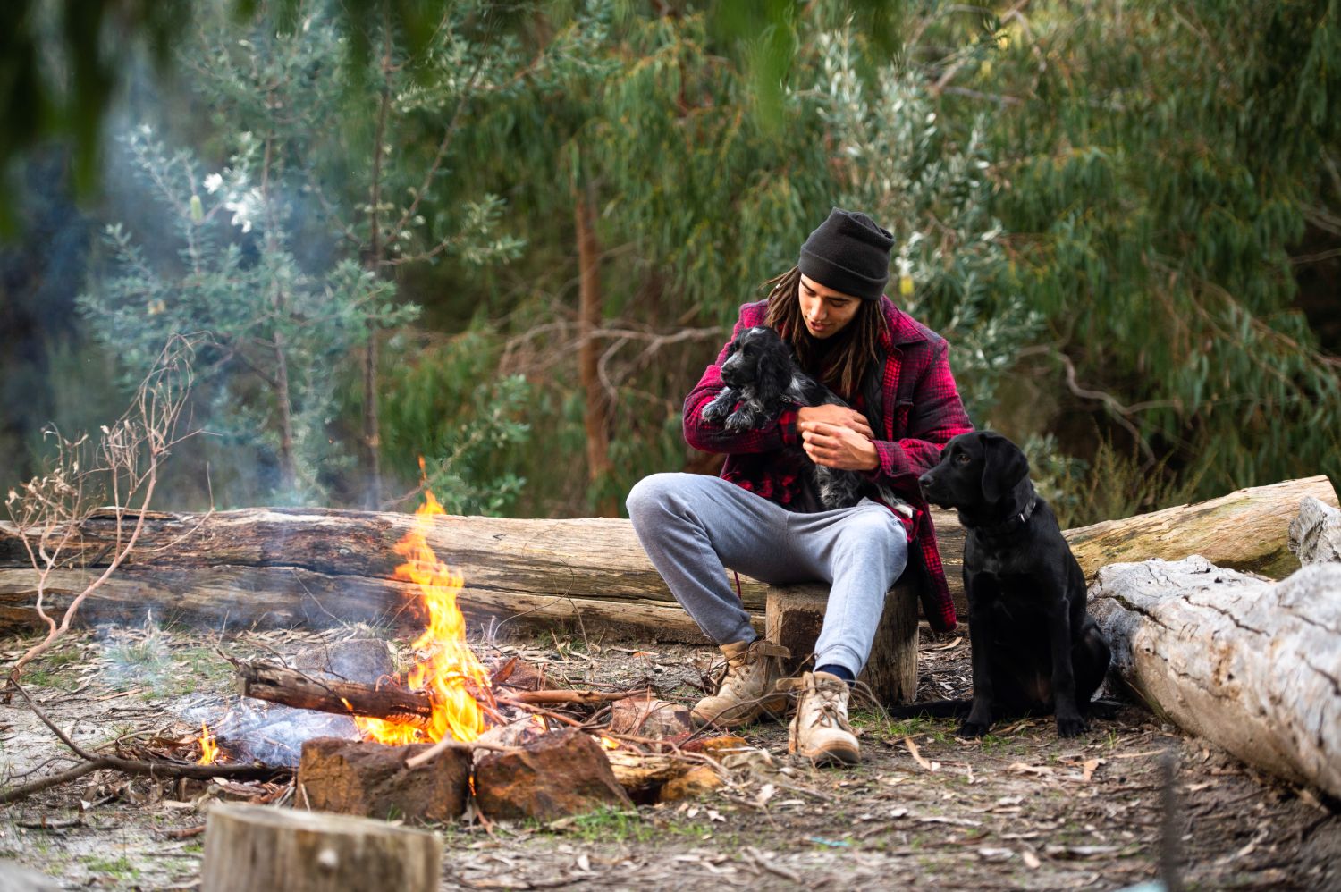 Man sits hugging his dog in the bush, by a burning fire